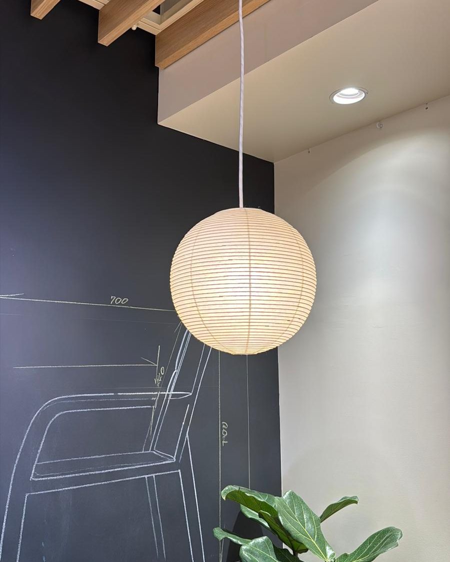 Isamu Noguchi AKARI 30A pendant lamp, lit, under timber beams beside a dark chalkboard wall: round natural white washi shade on bamboo ribbing, 30 cm across