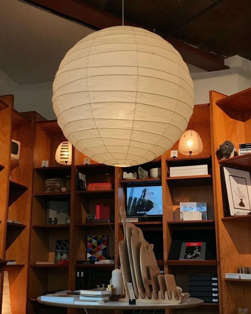 Isamu Noguchi AKARI 55D pendant lamp, lit, hanging over a room lined with wooden shelves: round natural white washi shade on bamboo ribbing, 55 cm across